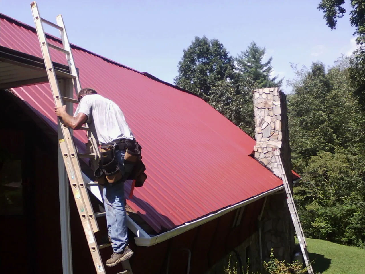Roofer on a ladder beside a red metal roof and stone chimney — Soffit & Fascia Repair by Roofing Brothers in Belmar